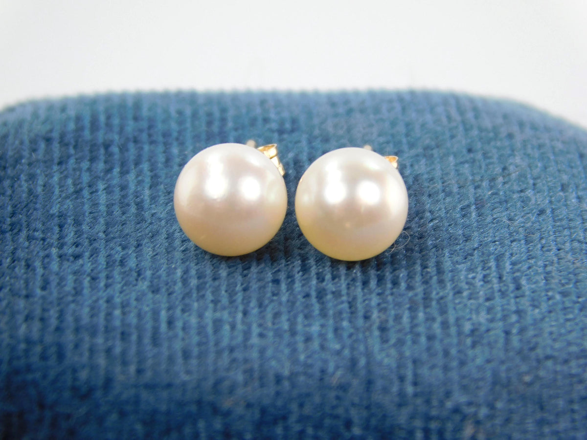 A close up of a pair of white cultured pearl stud earrings from the front. The yellow gold posts and backs aren't shown as much as the pearls themselves.  These are classic pearl studs.